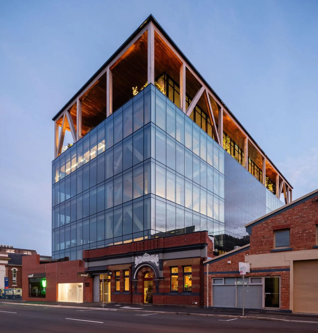St Luke&rsquo;s Health Headquarters at dusk: a glazed office volume with an exposed timber frame on the top floor, rising above a red-brick heritage street front